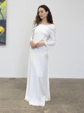 Woman wearing a fashion designer Adrian Hailwood  white off-shoulder dress standing against a plain wall.